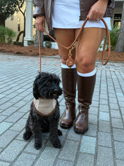 A small black dog wearing a beige and white striped bandana sits on a gray paved walkway while being held on a tan leash by a person wearing brown knee-high boots, a white skirt, and a brown jacket.