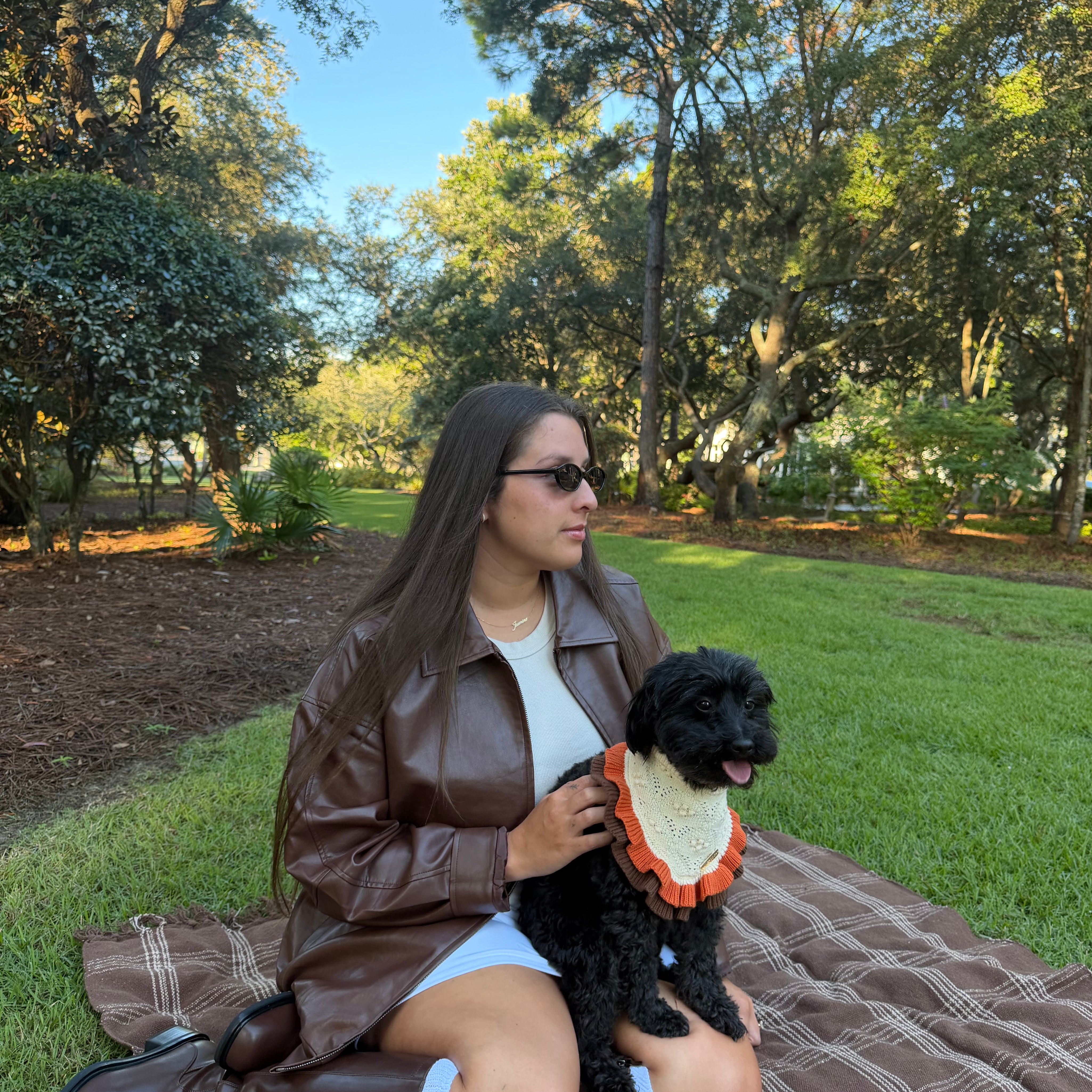 A woman sitting on a brown plaid blanket in a park with trees and grass, wearing a brown faux-leather jacket, white top, and brown knee-high boots, while holding a small black dog with an orange and cream ruffled collar.