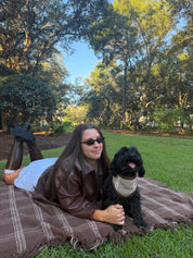 A woman in a brown leather jacket and sunglasses lies on a brown plaid blanket with her black dog in a sunny park setting with green grass and trees.