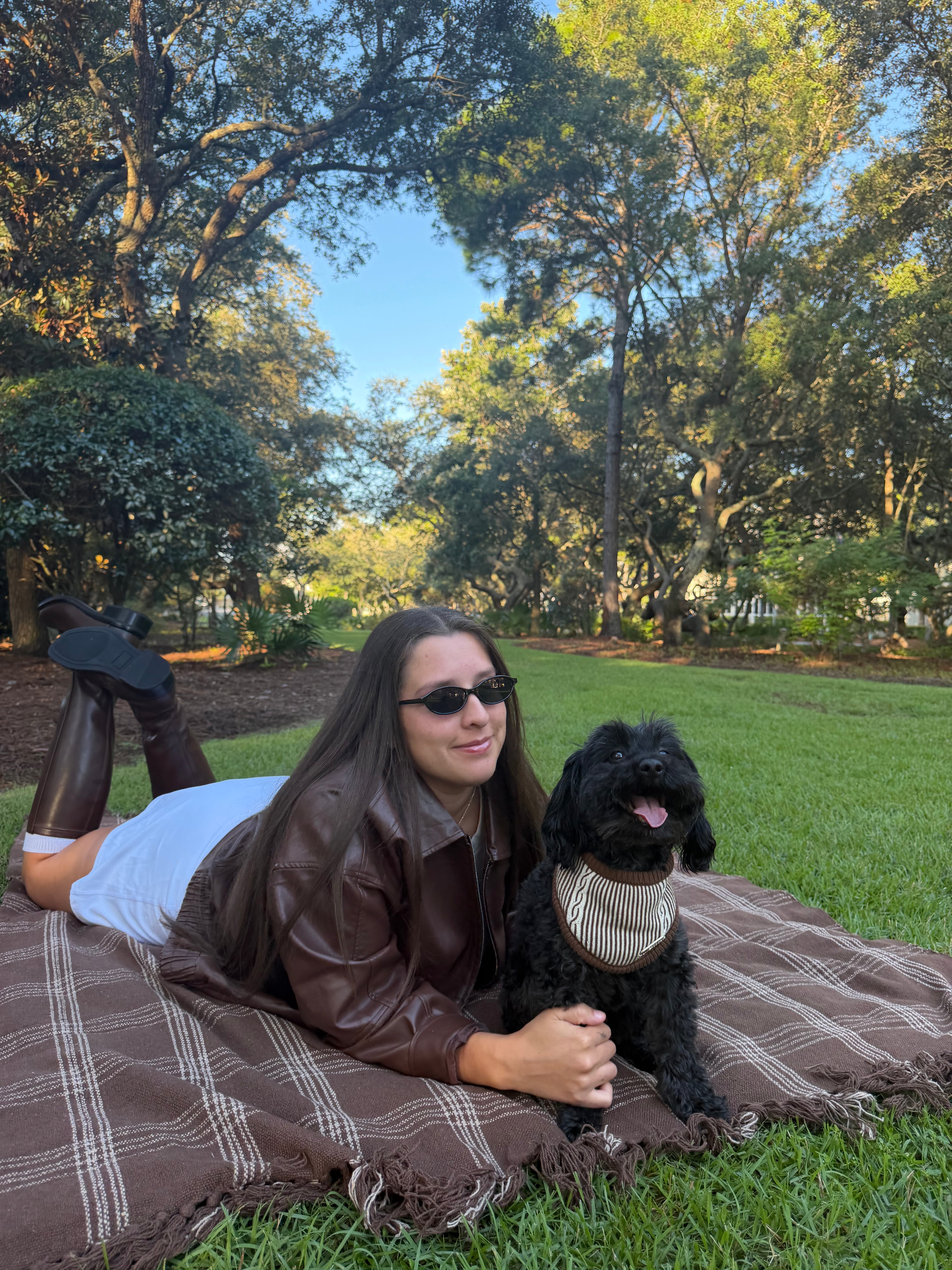 A woman in a brown leather jacket and sunglasses lies on a brown plaid blanket with her black dog in a sunny park setting with green grass and trees.