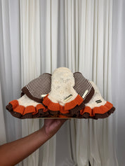 Three knit sun hats are held up by a hand against a white curtain background. The hats feature a cream knit crown with brown stripes on two, and a cream knit crown with an orange ruffled brim on all three.