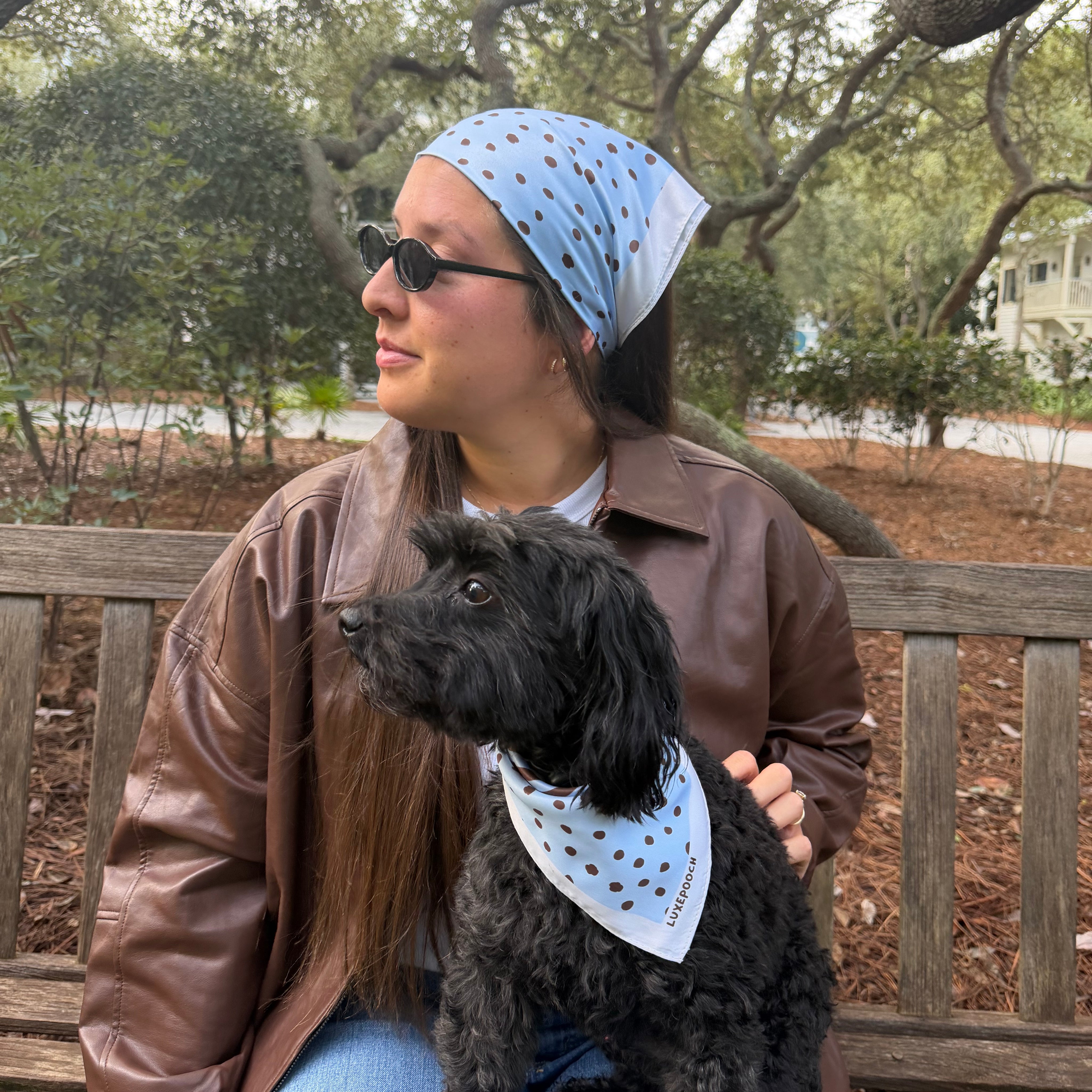 A woman sitting on a wooden park bench with a black dog, both wearing matching light blue bandanas with dark polka dots. The woman is wearing a brown leather jacket and blue jeans, and the background shows trees and a park setting.