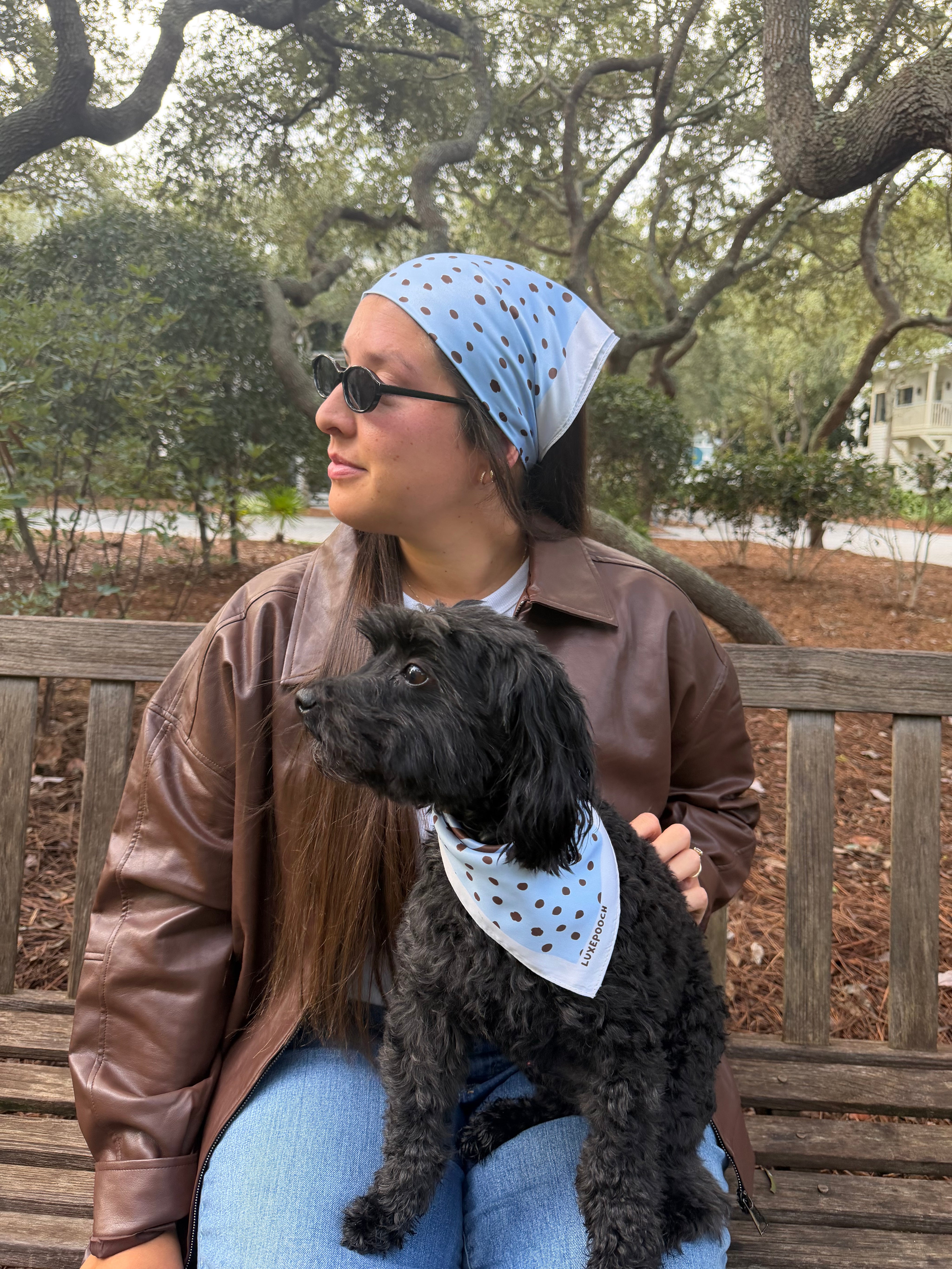 A woman sitting on a wooden park bench with a black dog, both wearing matching light blue bandanas with dark polka dots. The woman is wearing a brown leather jacket and blue jeans, and the background shows trees and a park setting.