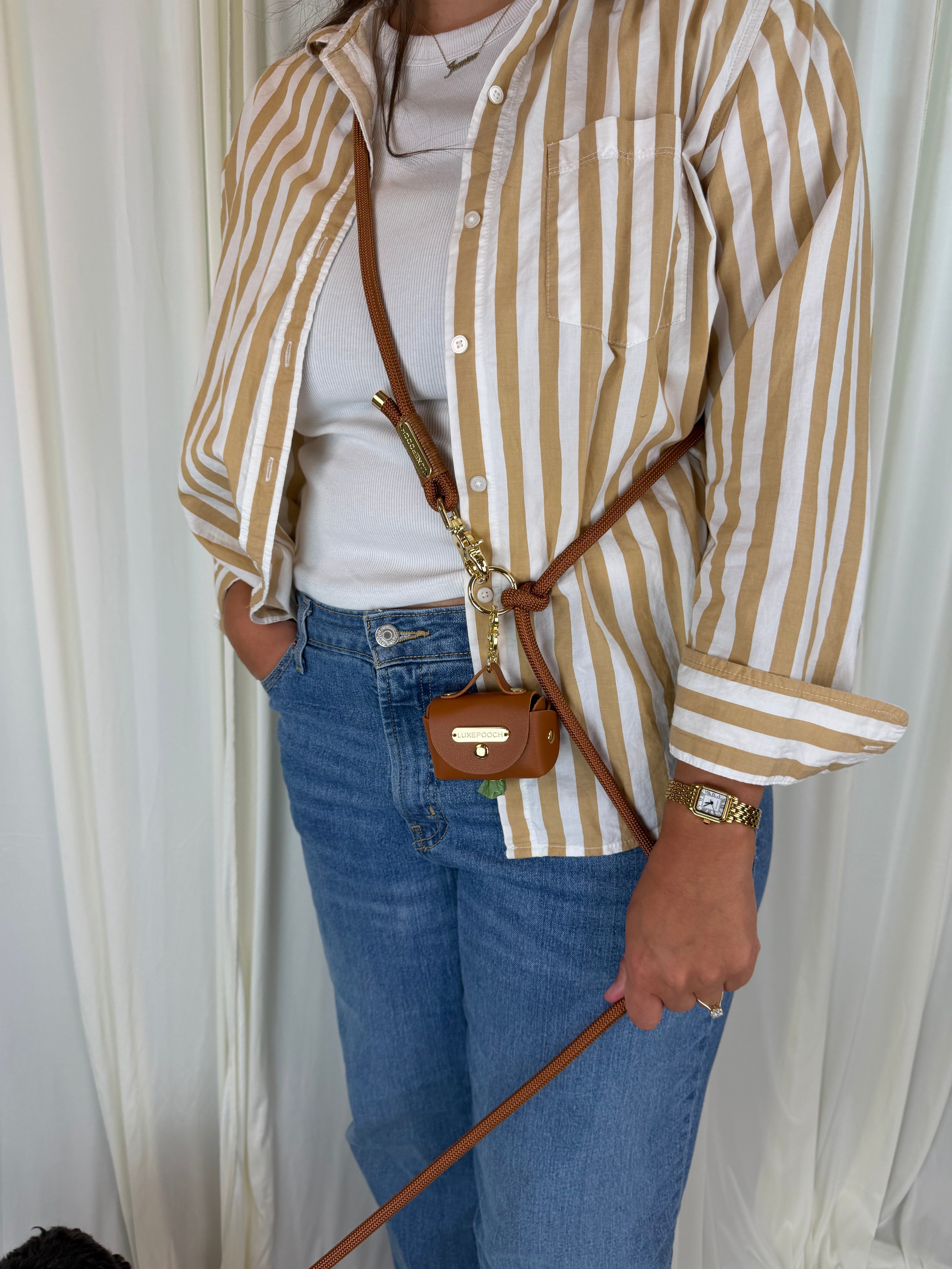 A person wearing a beige and white vertically striped button-up shirt, a white t-shirt, and blue jeans, carrying a small brown poop bag dispenser on a brown dog leash.