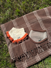 A brown plaid blanket with fringe is laid on grass, styled with two baby accessories: an off-white knit bib with orange ruffles and a striped knit cap.