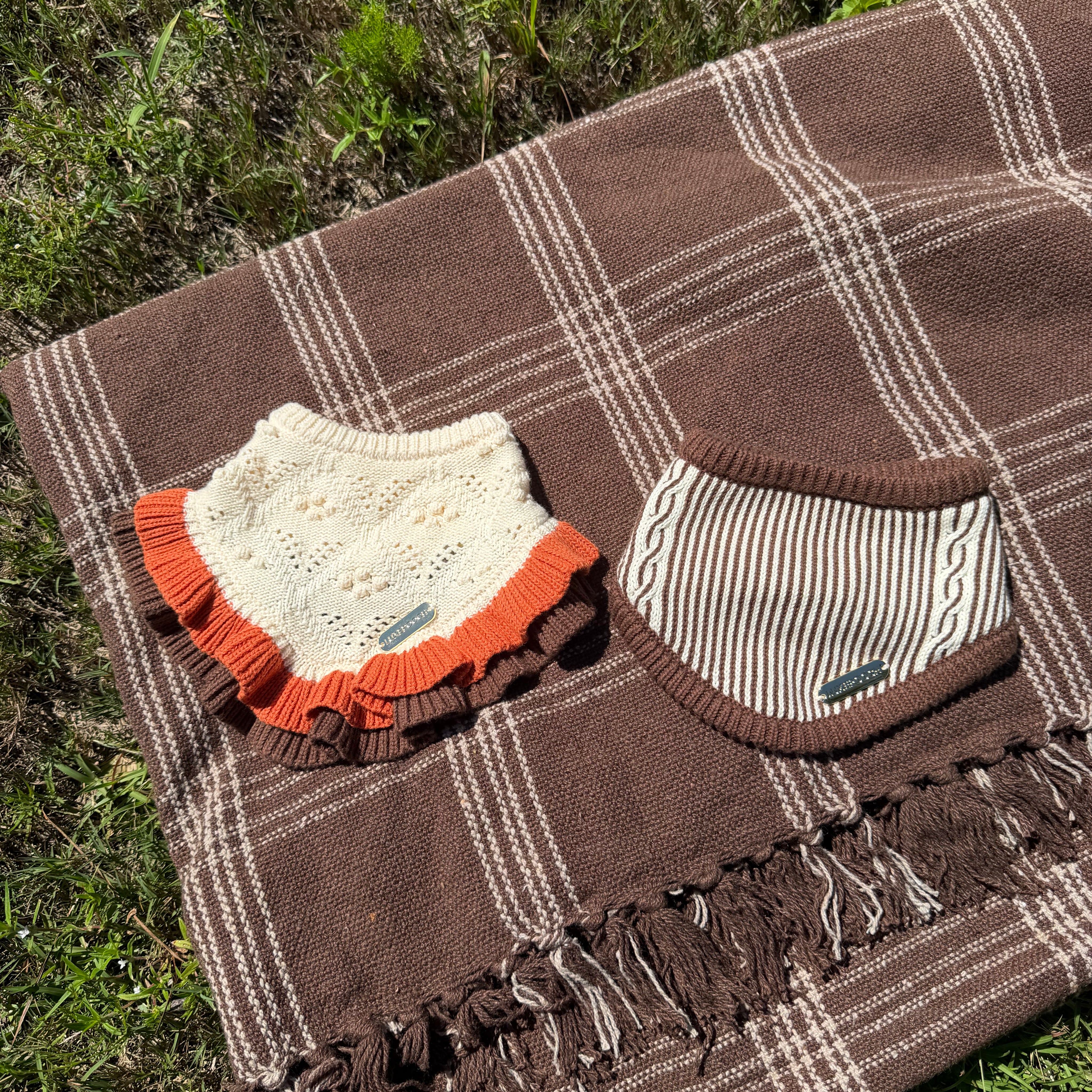 A brown plaid blanket with fringe is laid on grass, styled with two baby accessories: an off-white knit bib with orange ruffles and a striped knit cap.