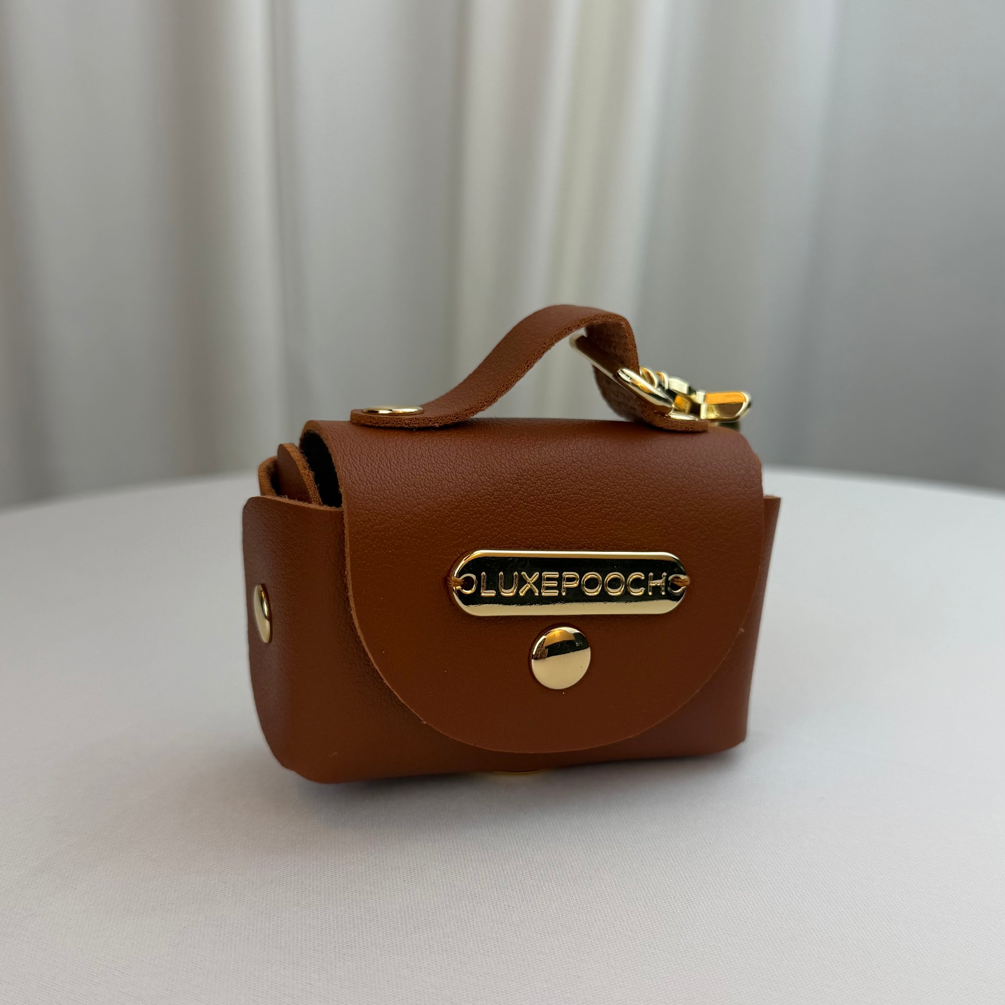 A small brown faux-leather dog waste bag dispenser with a top clasp and gold-tone hardware, featuring a metal plate on the front that reads 'LUXEPOOCH', set on a light gray surface with a soft gray curtain background.