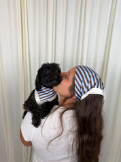 A woman and a small black dog are shown from the back, both wearing matching blue and white striped headscarves and a matching striped pet shirt. They are standing in front of a cream-colored curtain backdrop.
