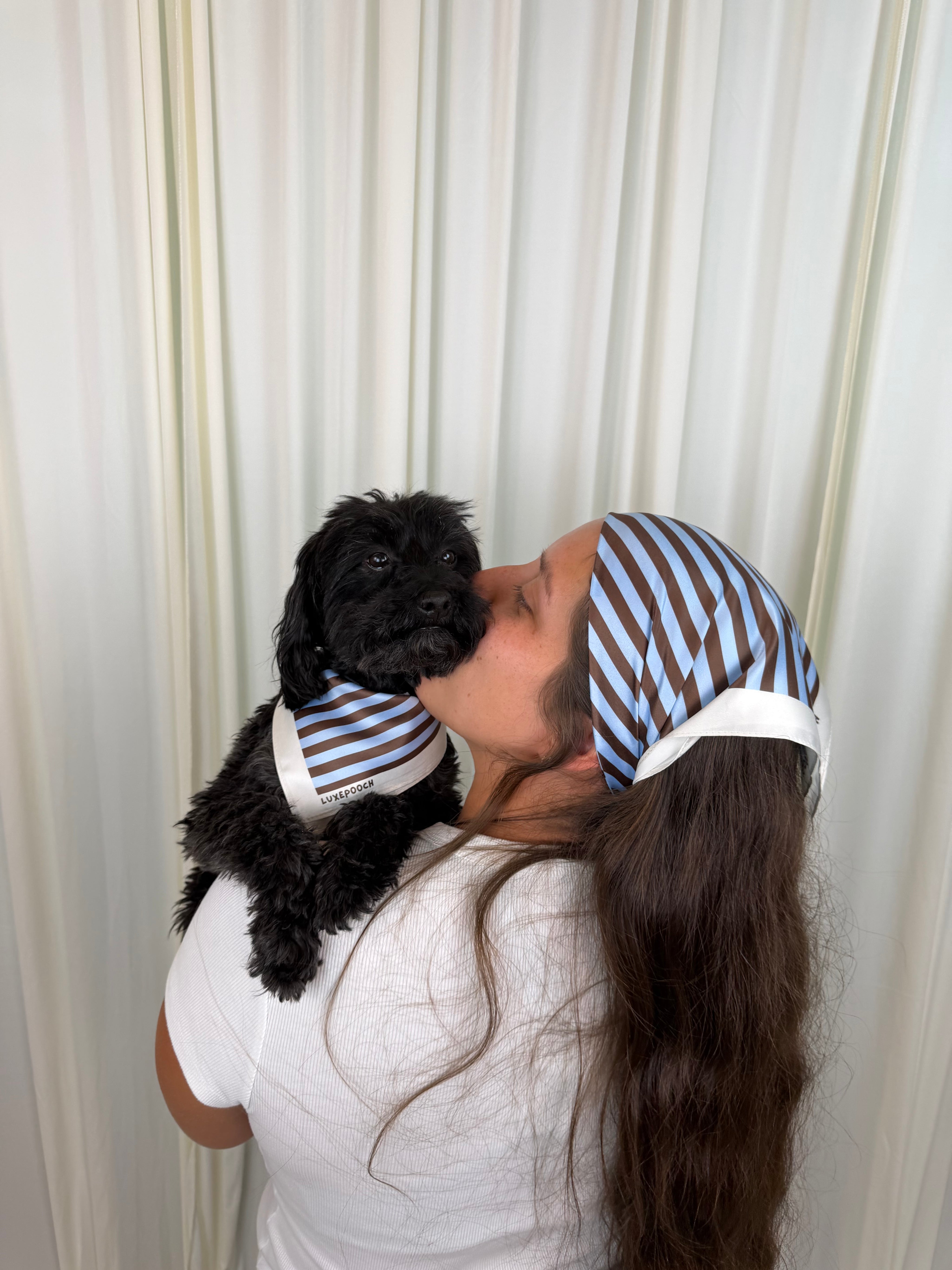 A woman and a small black dog are shown from the back, both wearing matching blue and white striped headscarves and a matching striped pet shirt. They are standing in front of a cream-colored curtain backdrop.