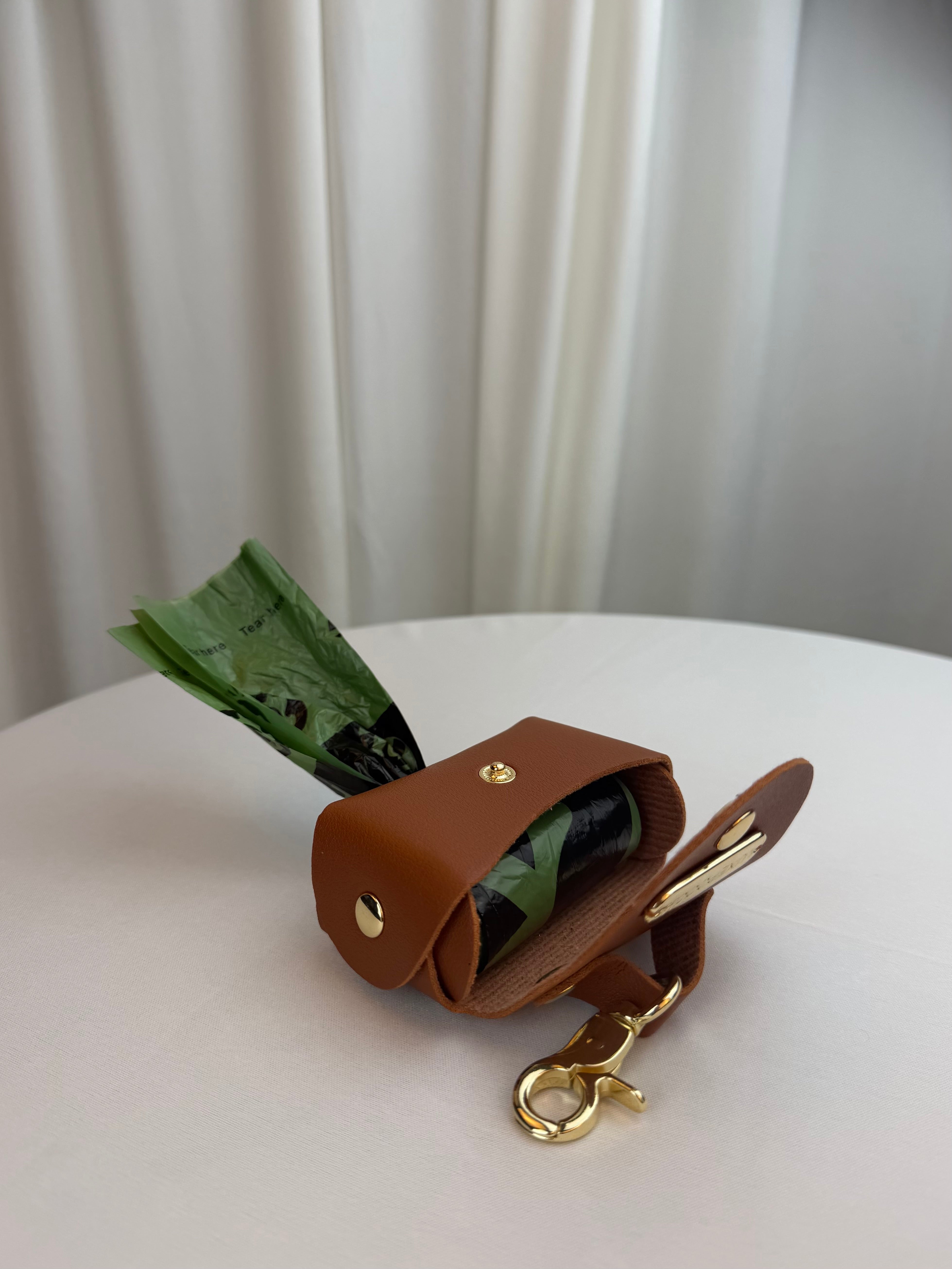 A brown leather dog poop bag holder with gold-tone hardware, shown open with a green bag of dog poop inside, resting on a white surface with a soft-focus curtain background.