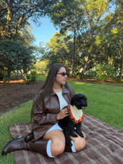 A woman sitting on a brown plaid blanket in a park with trees and grass, wearing a brown faux-leather jacket, white top, and brown knee-high boots, while holding a small black dog with an orange and cream ruffled bandana.