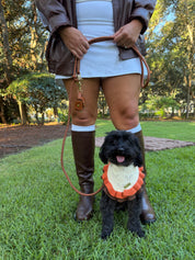 A black dog wearing a cream and orange knit bandana sits on a green lawn while a person in a brown jacket and boots holds its leash.