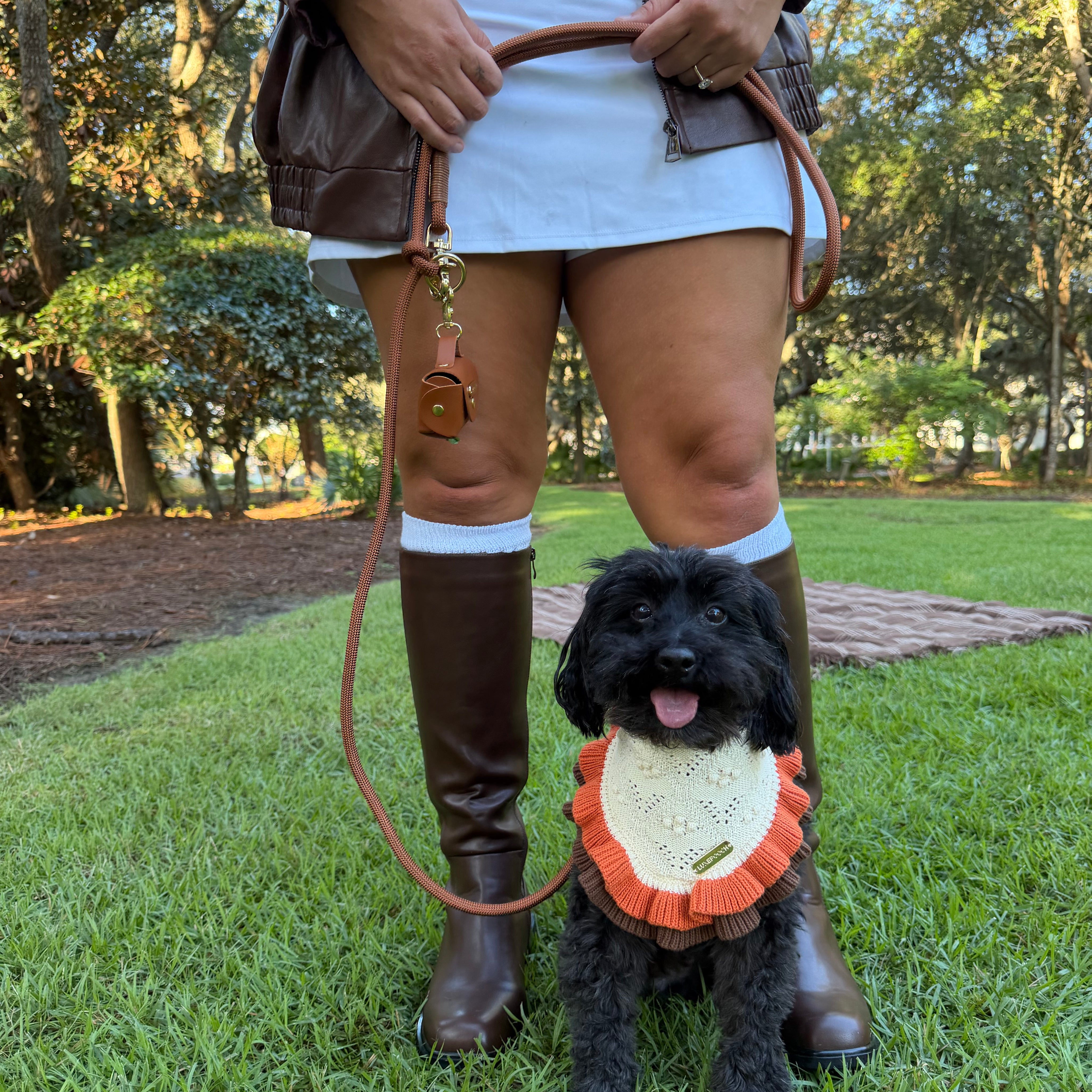 A black dog wearing a cream and orange knit bandana sits on a green lawn while a person in a brown jacket and boots holds its leash.