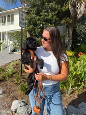 A woman wearing sunglasses, a white t-shirt, and blue jeans holds a small black dog wearing a brown leather shoulder bag outdoors in front of a house and greenery.