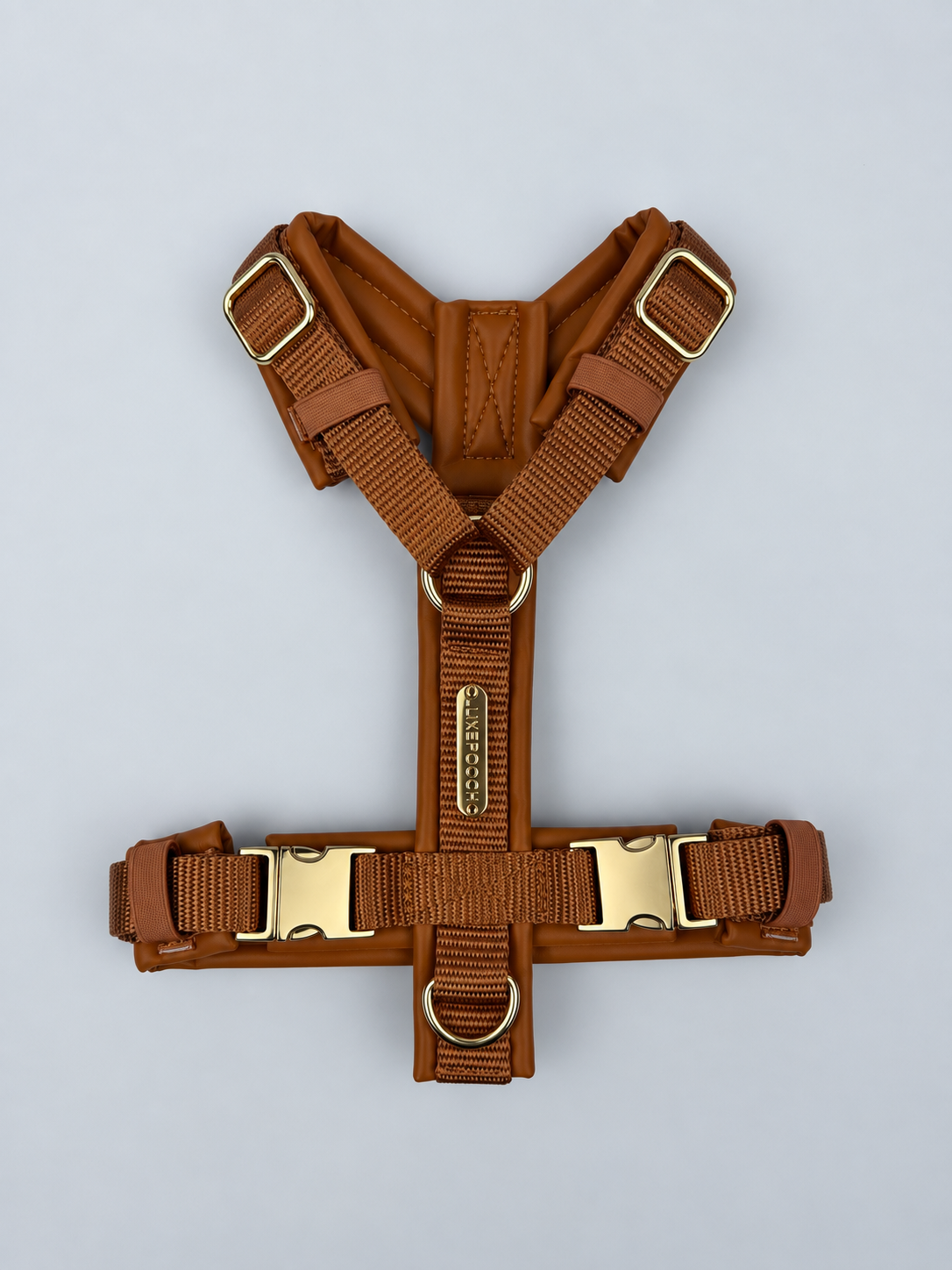 A brown dog harness with adjustable webbing straps and gold-tone metal hardware, laid flat on a light gray background.