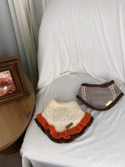 Two knitted pet bandanas displayed on a white-covered chair next to a wooden side table with a framed picture and an orange pumpkin.